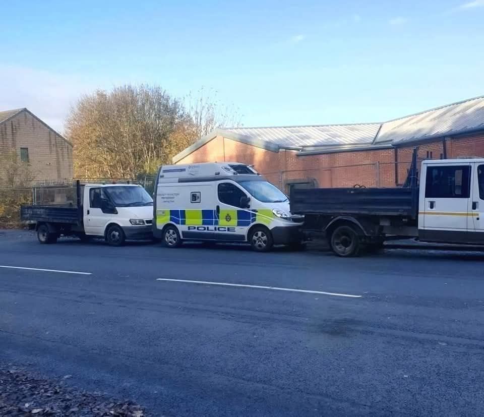 Policajné auto medzi dvoma nákladnými vozidlami na parkovisku pred priemyselnou budovou.