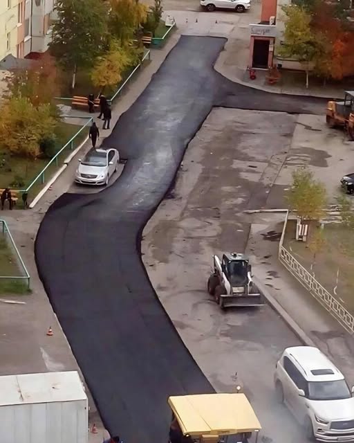 Cesta s čerstvým asfaltom v mestskom prostredí. Pár áut jazdí po novom povrchu, obklopené jesennou zeleňou.