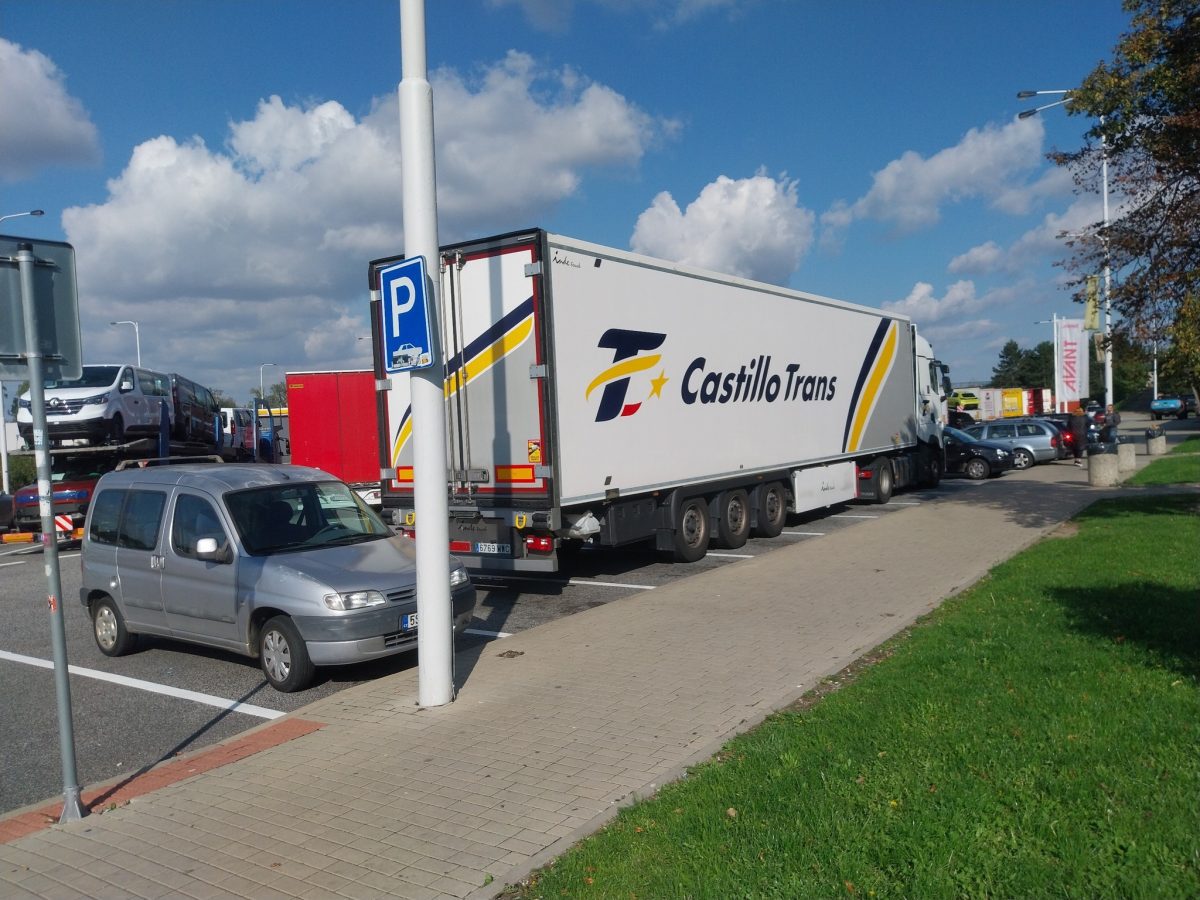 Kamión na parkovisku vedľa osobného auta a odťahovej plošiny, pod modrou oblohou s oblakmi.