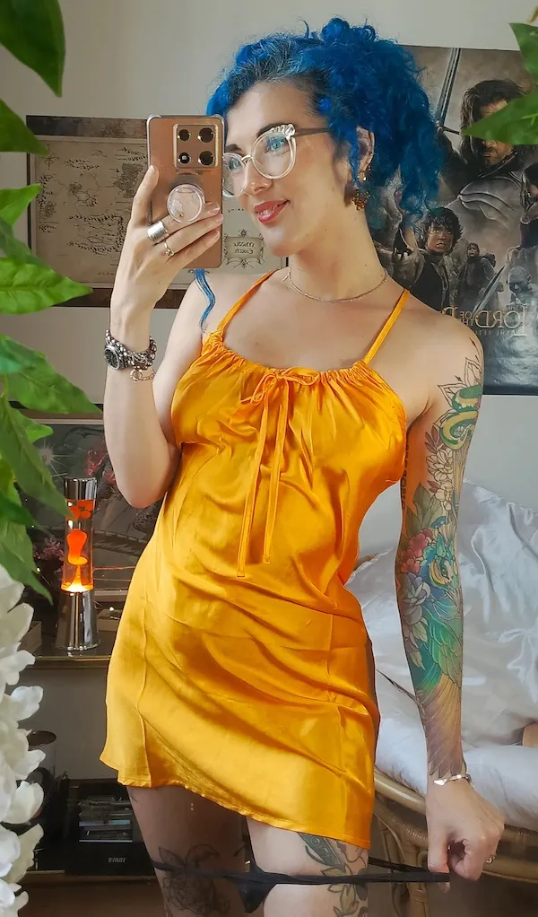 Osoba so modrými vlasmi a okulármi v oranžových šatách fotí selfie v izbe plnej rastlín a plagátov.