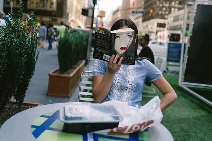 Žena na ulici číta knihu so zladeným obalom tváre, okoloidúci mestský život v pozadí.