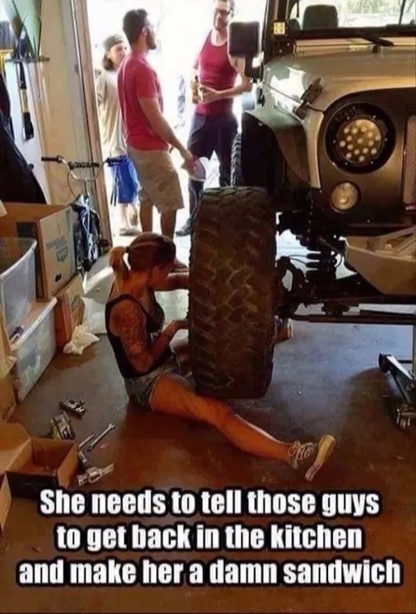 Žena opravuje auto v garáži, zatiaľ čo muži stoja bokom a rozprávajú sa.