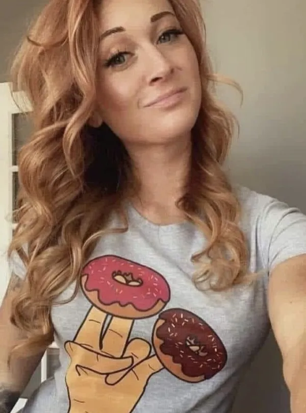 Žena s dlhými ryšavými vlasmi v tričku s motívom donútov, usmievajúc sa do kamery v svetlej miestnosti.