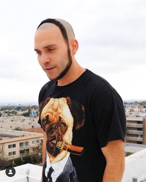 Muž s originálnou účesovou variáciou, oblečený v tričku s potlačou mopsa s cigárou, na streche mestského domu.