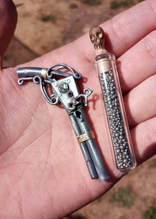 Miniatúrna kovová pištoľ s ornamentom a sklená fľaštička s guľôčkami na dlani.