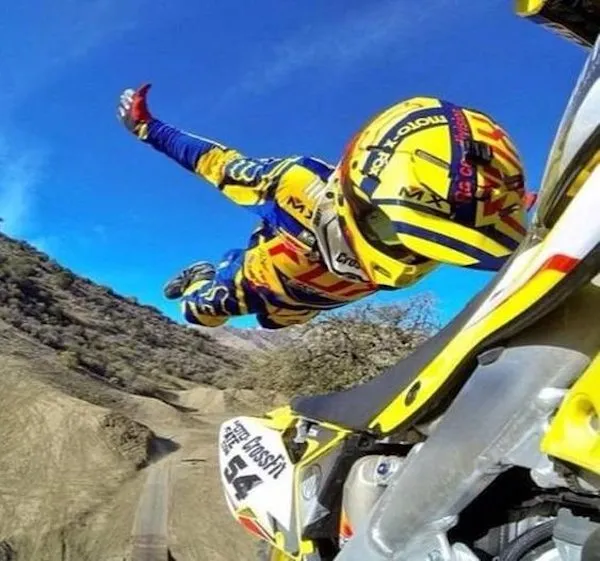 Motokrosový jazdec vo farebnej prilbe vo vzduchu nad prašnou dráhou pod modrou oblohou.