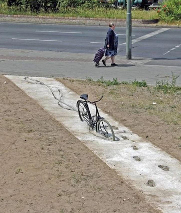 Bicykel zapadnutý v betóne, vedľa chodníka prechodí žena s nákupnou taškou na kolieskach.