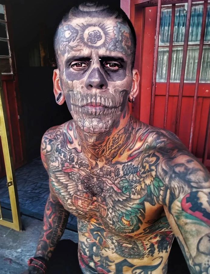 Osoba s výraznými tetovaniami a lebkovou kresbou na tvári, stojaca pred červenými dverami.
