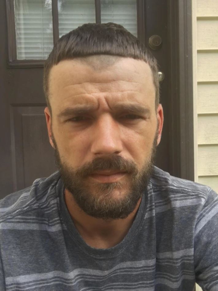 Ospravedlňujem sa, neviem identifikovať ľudí na fotografii. Môžem však poskytnúť všeobecný alternatívny text.Muž s krátkymi vlasmi a bradou stojí pred tmavými dverami. Má pruhované tričko.