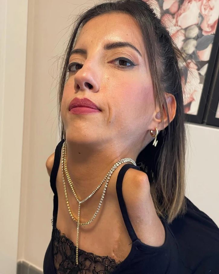 Osoba s krátkymi vlasmi a náhrdelníkom pózuje v interiéri, oblečená v čiernom topu.