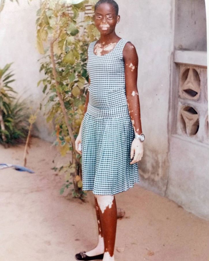 Osoba v károvaných šatách stojí vonku, obklopená rastlinami, na stenách vidno náznaky starnutia.