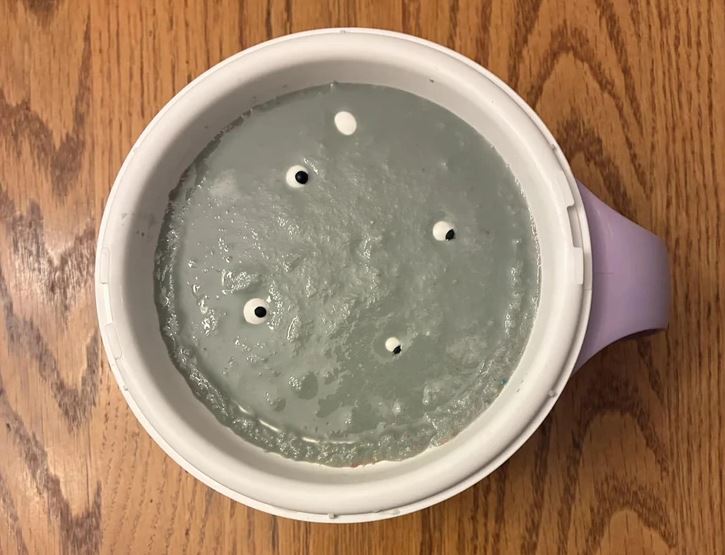 Uzavretá plastová nádoba s otvormi na povrchu šedej hmoty, položená na drevenom stole.