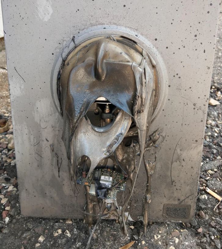 Roztavený elektronický panel v dôsledku prehriatia, s odkrytou vnútornou elektronikou na pozadí betónu.