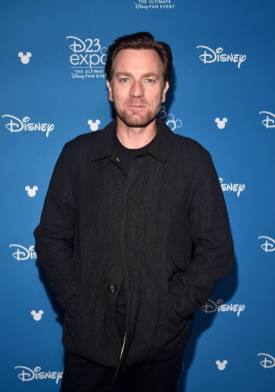 Osoba v čiernej bunde pózuje na modrom pozadí s logami Disney a D23 Expo.