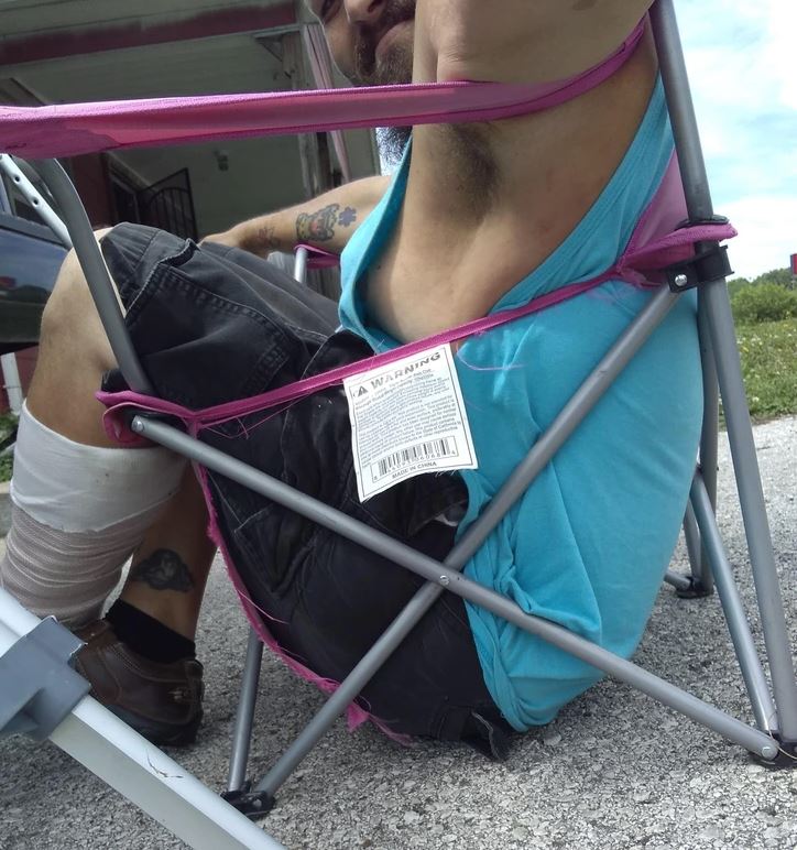 Osoba prehnutá v skladacej stoličke na dvore, modré tričko, s tetovaním na nohe a obviazaným kolenom.