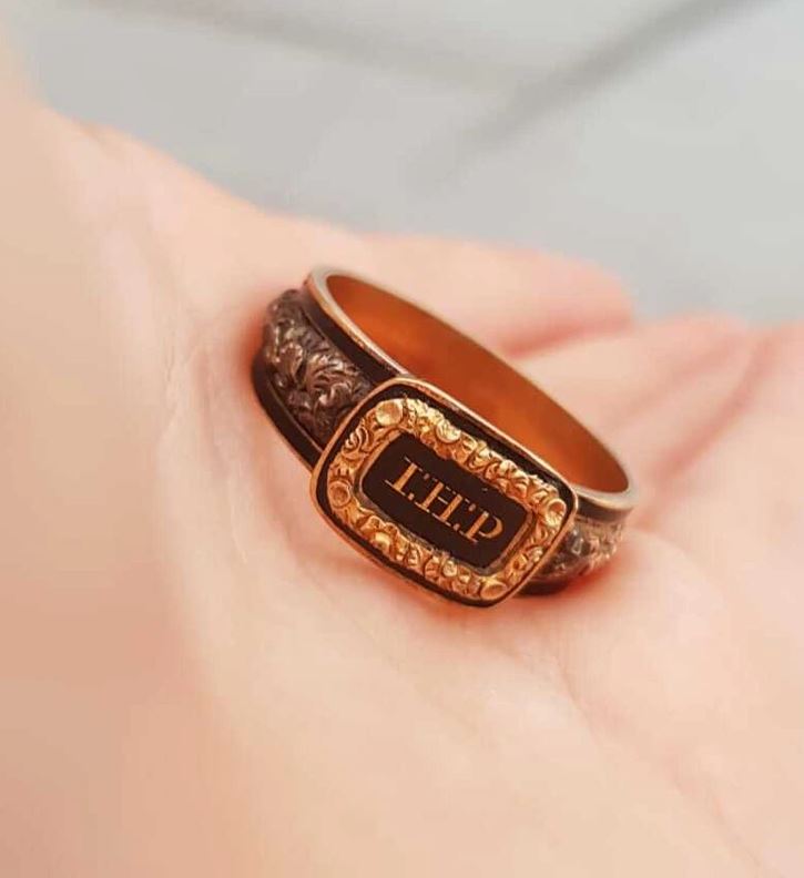 Zlatý prsteň s vyrytými iniciálmi „T.H.P.“ v ozdobnom ráme, položený na dlani.