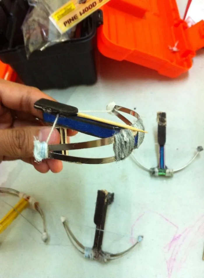 Ručne vyrobený miniatúrny kuša z kovu a dreva na bielom pozadí s oranžovým boxom v pozadí.