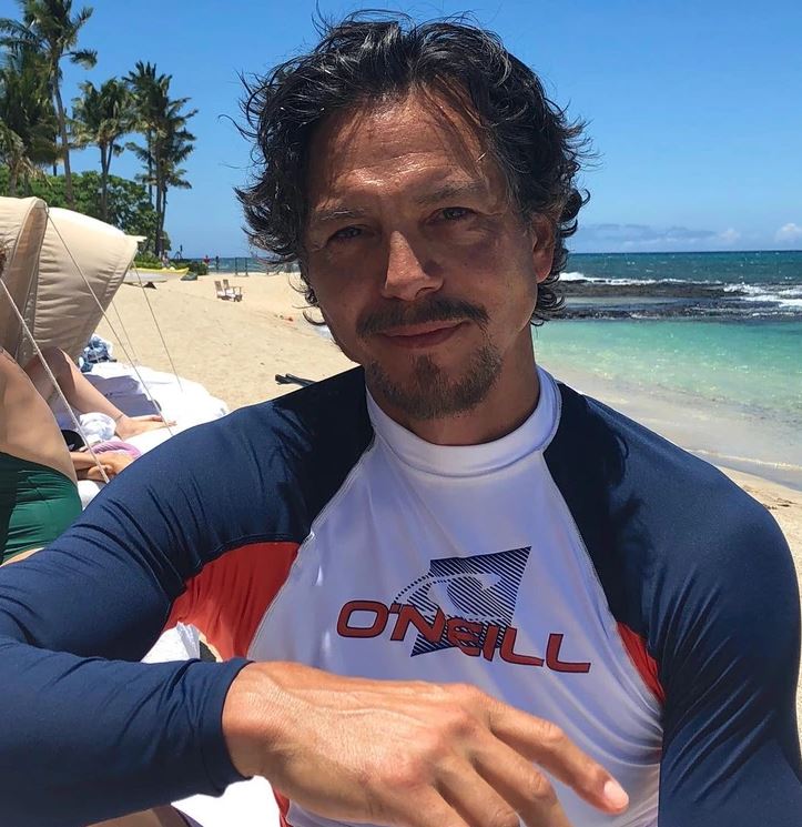 Osoba na pláži v slnečnom počasí s modro-bielym športovým tričkom, ležérne pózuje pri mori.