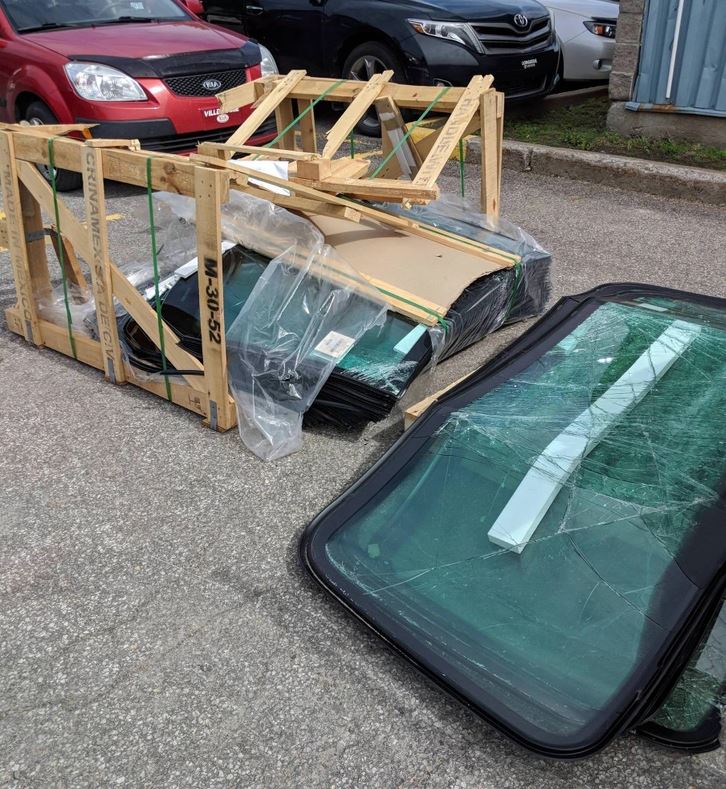 Rozbité čelné sklá vo drevenom ráme na parkovisku, okolo stojí viacero áut.