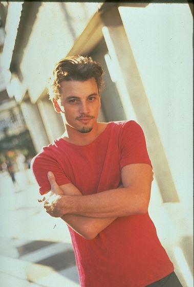 Muž v červenom tričku stojí na ulici, opiera sa o stenu a pozerá do kamery.