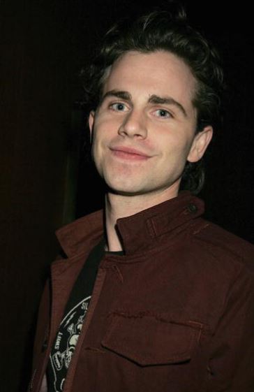 Osoba v hnedé bunde sa usmieva do objektívu na tmavom pozadí.