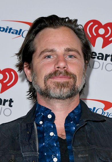 Osoba s bradou na červenom koberci pred logom iHeartRadio a CapitalOne, oblečená v rifľovej bunde.