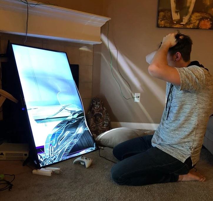Osoba v panike pred rozbitou obrazovkou televízora, sedí na koberci s videohrou a ovládačom.
