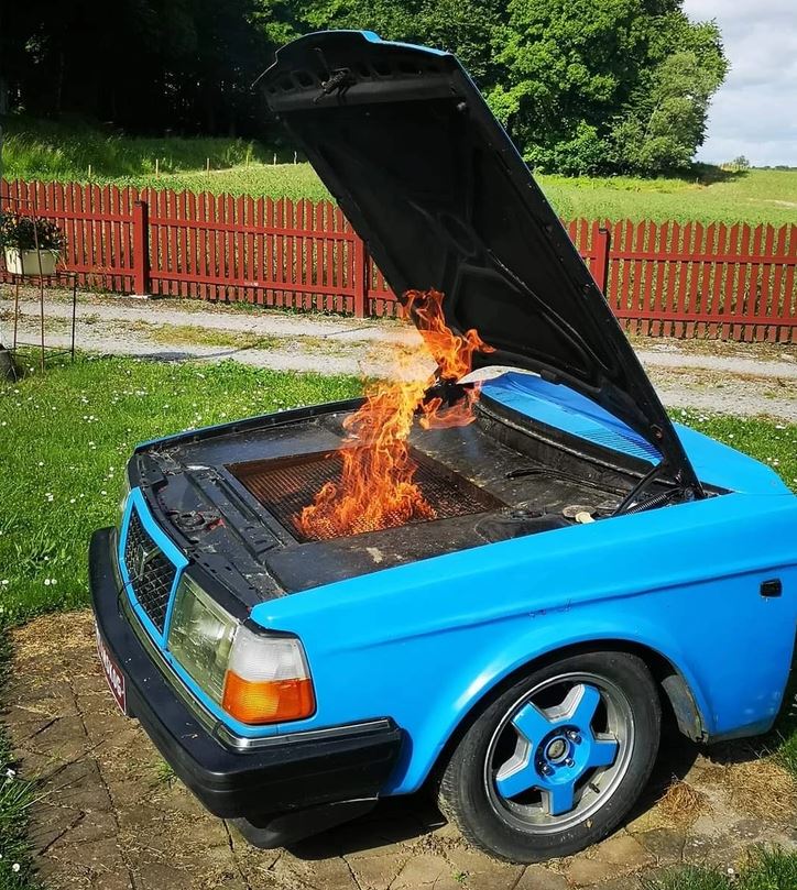 Auto s otvorenou kapotou, používané ako vonkajší gril s plameňmi, v záhrade pri červenom plote.