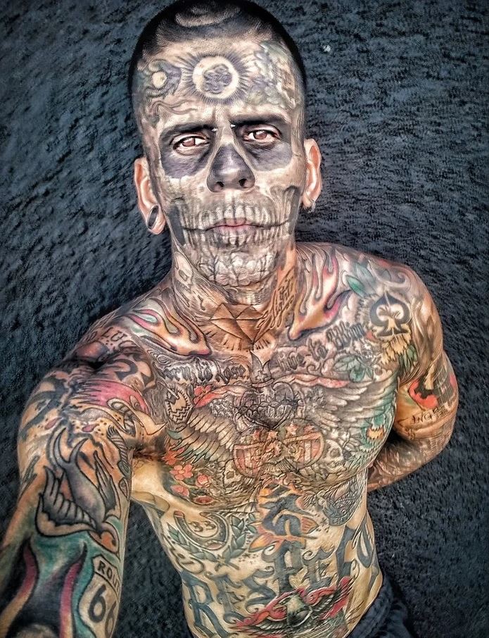 Osoba s výraznými tetovaniami a lebkovým motívom na tvári, pozadie je tmavé a kontrastné.