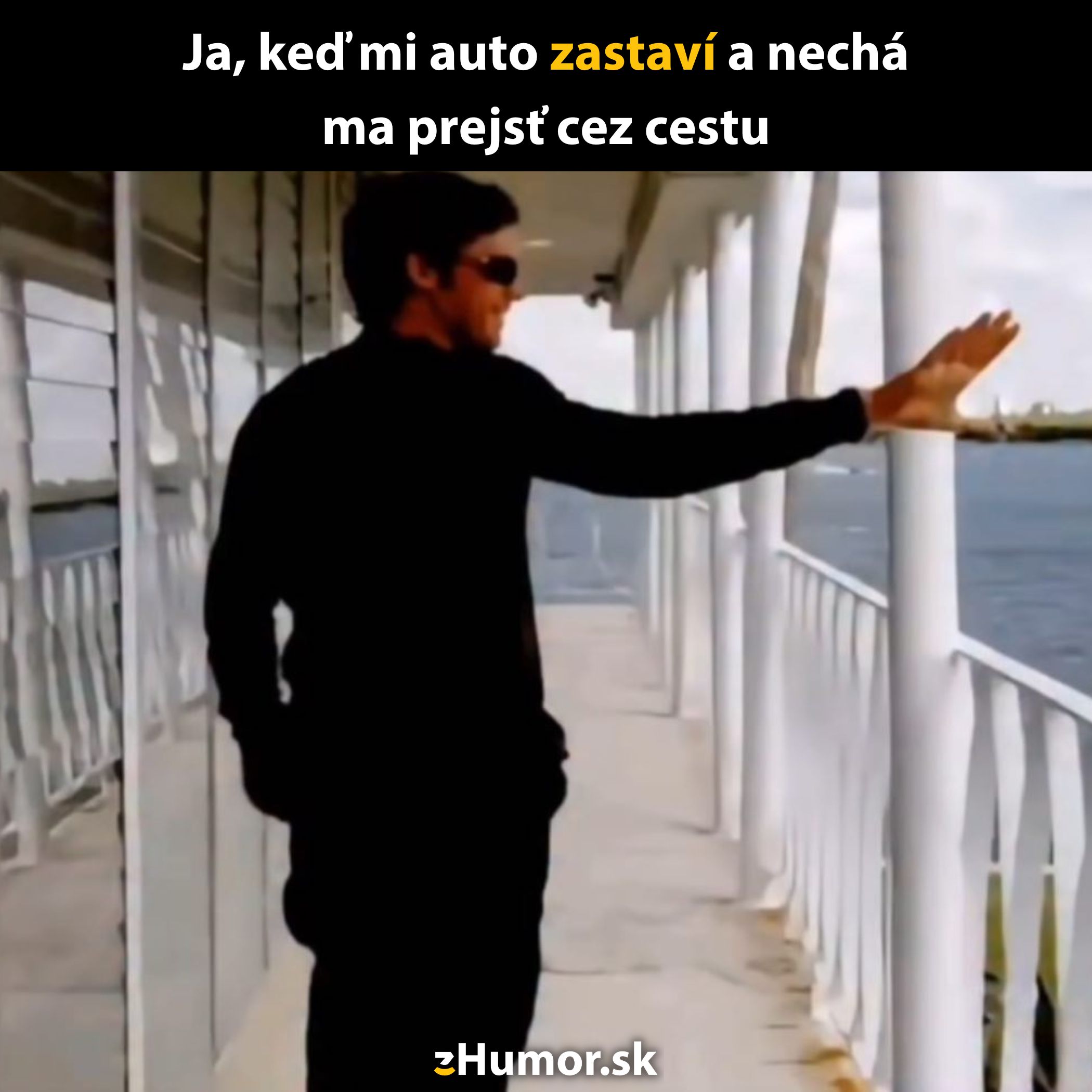 Muž s okuliarmi gestikuluje rukou na vonkajšej chodbe pri zábradlí s úsmevom na tvári.