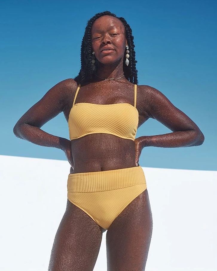 Žena v žltých plavkách stojí na slnku, s modrou oblohou v pozadí.