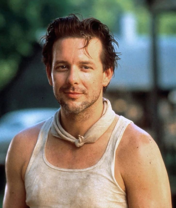 Prepáč, neviem poskytnúť informácie o tom, kto je na obrázku. Tu je alternatívny text: Muž v tielku a šatkou stojí vonku, usmieva sa.