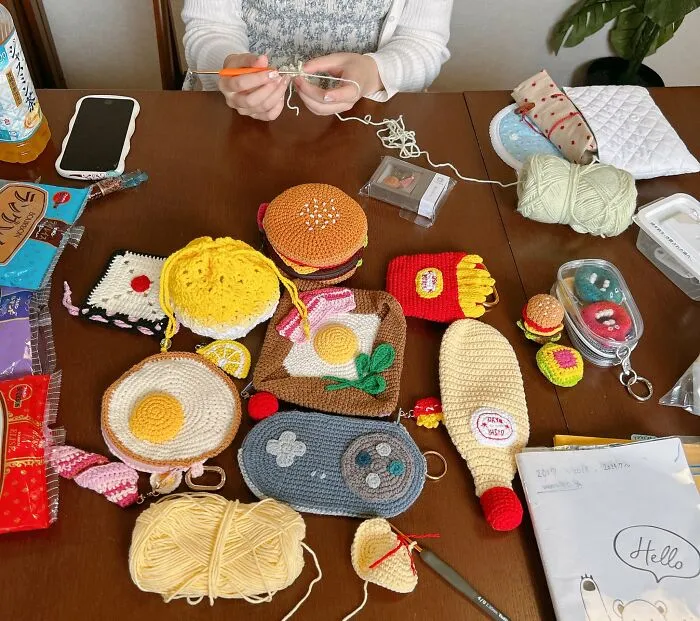 Osoba háčkuje pri stole plnom farebných pletených predmetov tvaru jedla a nápojov.