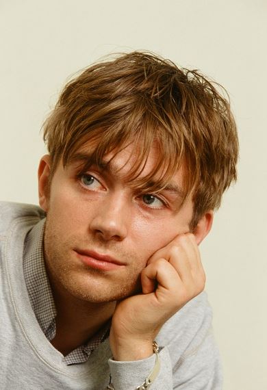 Mladý muž s blond vlasmi zamyslene podopiera tvár rukou, oblečený v šedom svetri.