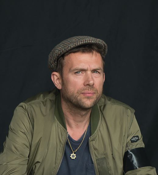 Osoba v zelenej bunde a čiapke pozujúca pred tmavým pozadím.