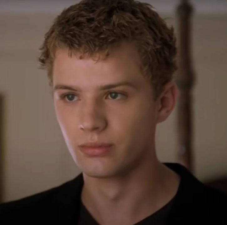 Osoba so svetlými vlasmi a sústredeným výrazom v interiéri, oblečená v tmavom odeve.