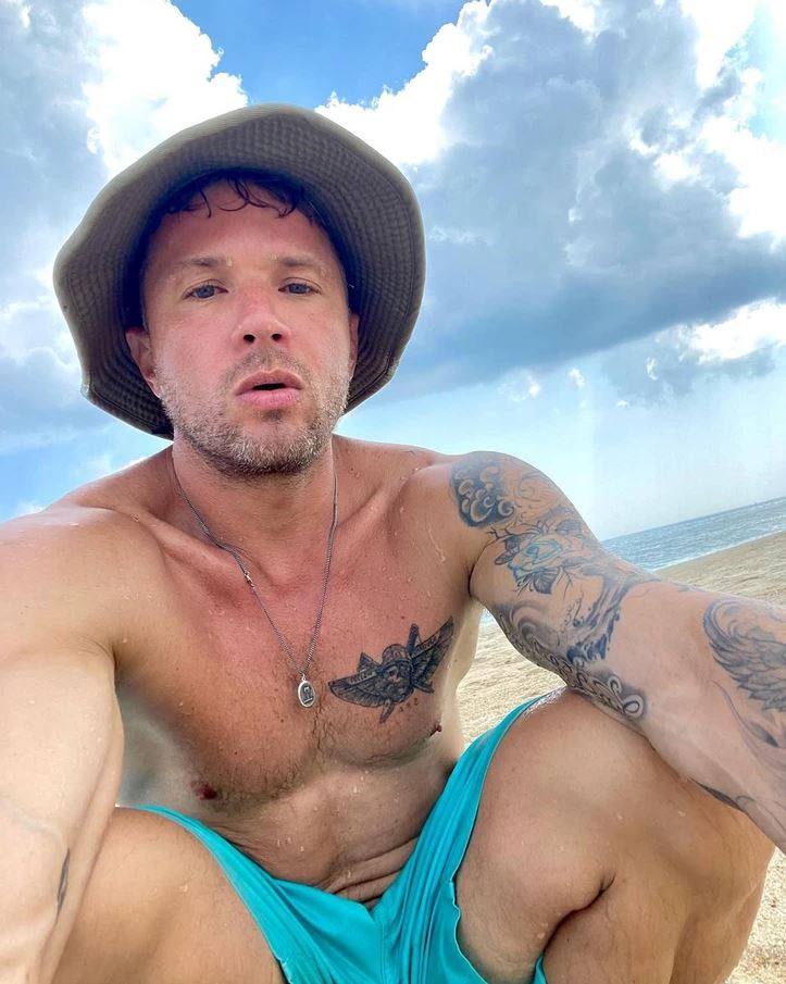 Muž v klobúku na pláži s tetovaním, obloha s mrakmi v pozadí.