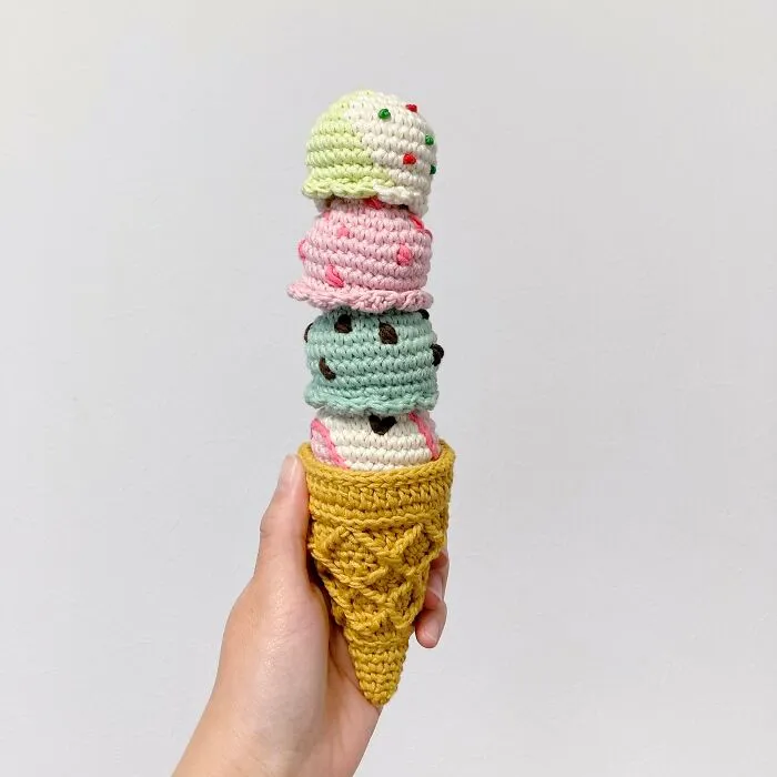 Háčkovaný zmrzlinový kornútok s rôznofarebnými kopčekmi, držiaci v ruke, na bielom pozadí.