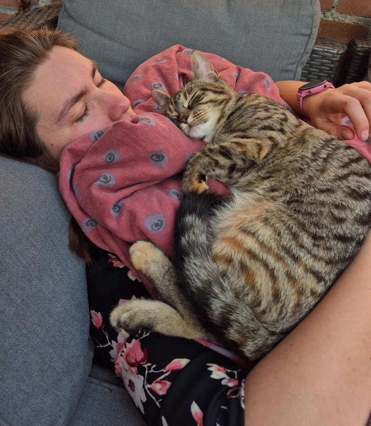 Osoba so zatvorenými očami leží na gauči, objímajúc spiacu mačku na červenej deke.