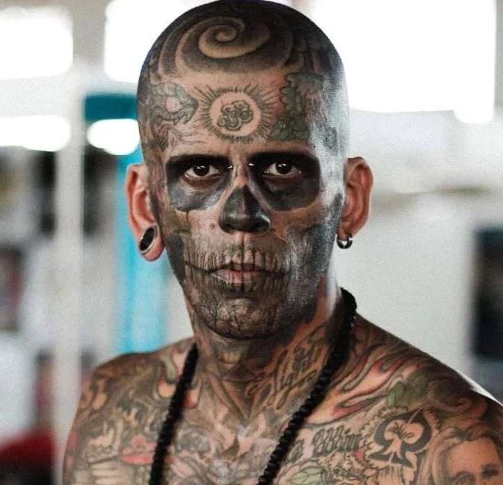 Osoba s tetovaniami na tvári a tele, evokujúca vzhľad lebky, pózuje v interiéri.