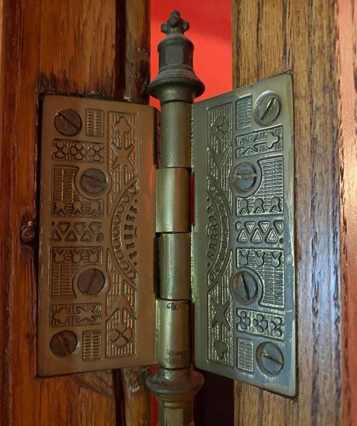 Ornamentálny kovový pánt na dverách s vyrezávanými vzormi a dreveným rámom na pozadí.