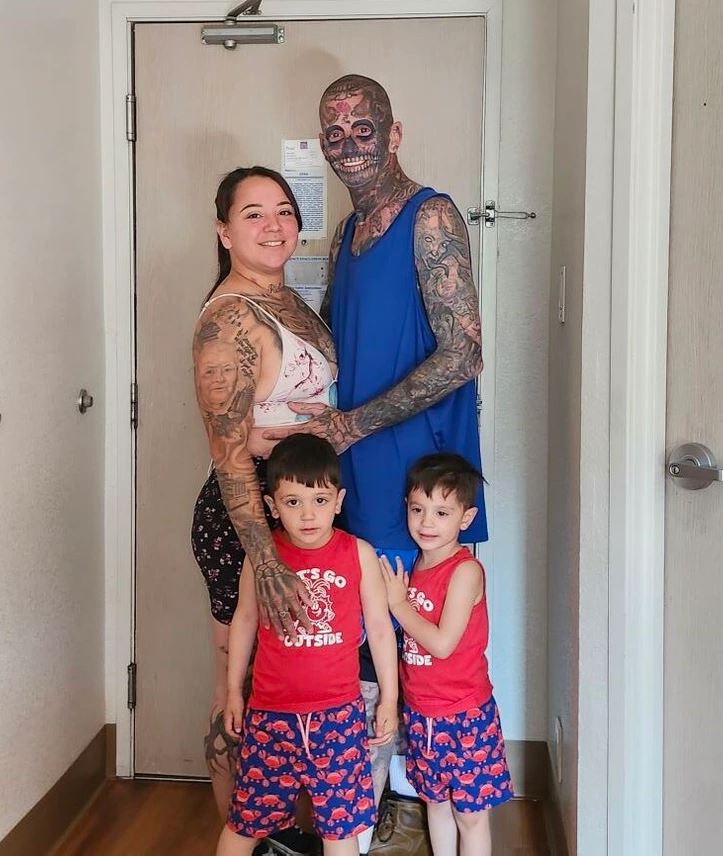 Ľudia s tetovaniami stoja spolu pred dverami, vrátane dvoch detí v červených outfitoch.