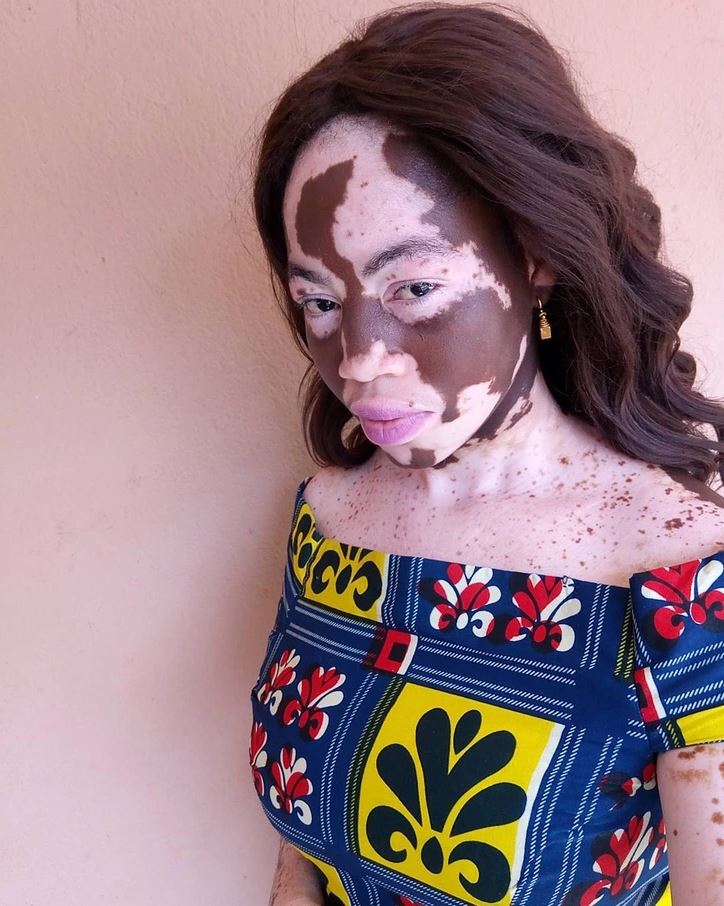 Žena s výrazným vzorom na tvári oblečená v pestrofarebných šatách pózuje pred béžovou stenou.