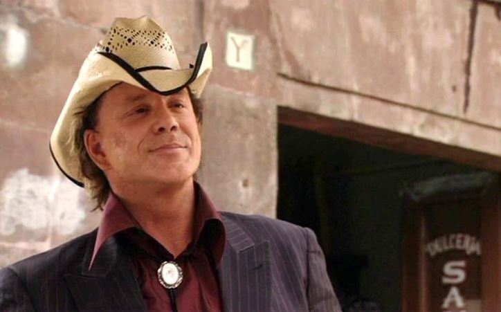 Muž v klobúku pred starou budovou, v elegantnom saku a košeli s doplnkom kravaty na šnúrke.