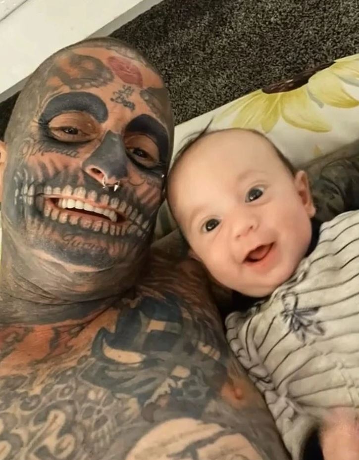 Otec s tetovaním a jeho usmiate bábätko na podložke s kvetinovým vzorom.