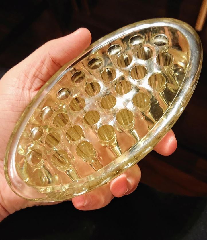 Priehľadný plastový predmet s otvormi držiaci v ruke, použiteľný ako masážny nástroj alebo podložka.