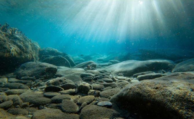 Podvodný scenár s lúčmi svetla osvetľujúcimi kamenné dno pod priezračnou modrou vodou.