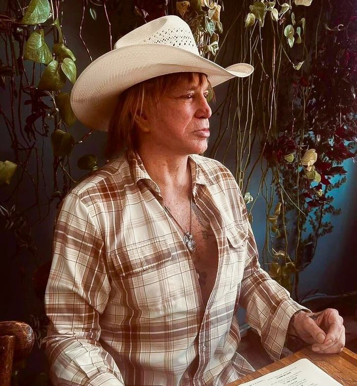 Osoba v kovbojskom klobúku sedí pri stole, obklopená visiacimi rastlinami.