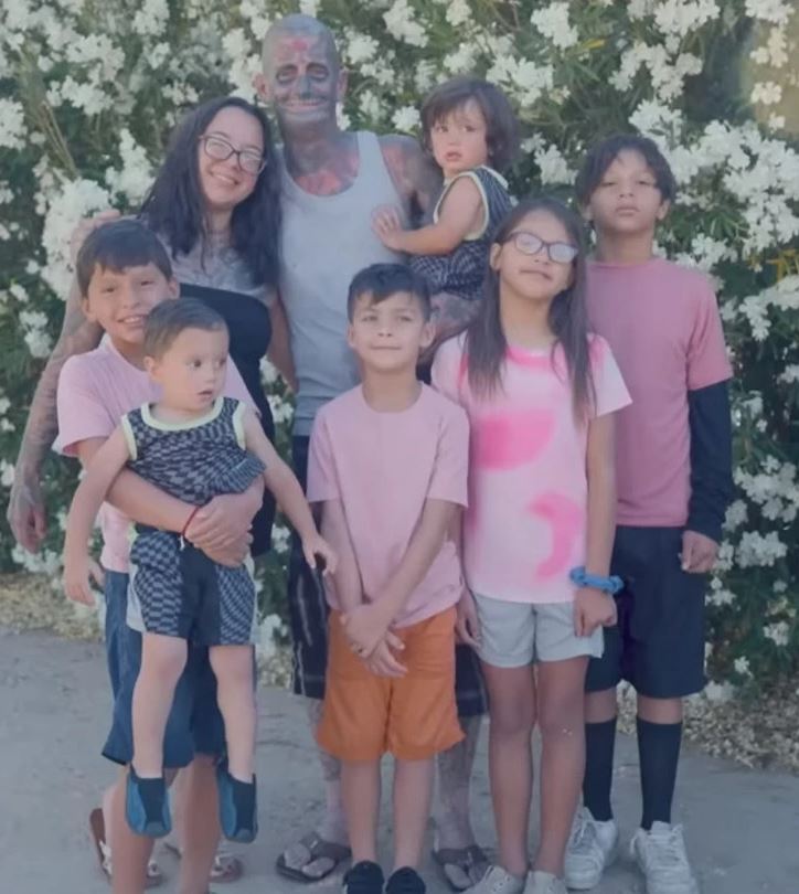 Rodina stojí pred kvitnúcimi bielymi kríkmi, oblečení v neformálnych ružových a šedo-modrých odevoch.