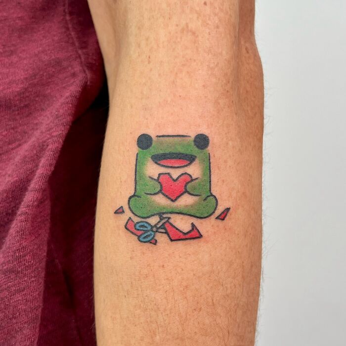 Tetovanie žaby s červeným srdcom a nožnicami na ruke.